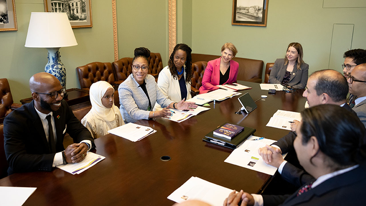 Photo of a diverse group of people in professional attire smiling and seated at a long table, sitting among adults is one child in a white headscarf.