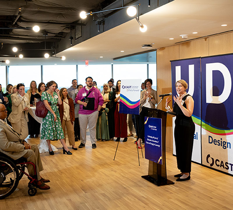 Photo of CAST CEO Lindsay Jones speaking to event guests, standing in front of three banners spelling out UDL.