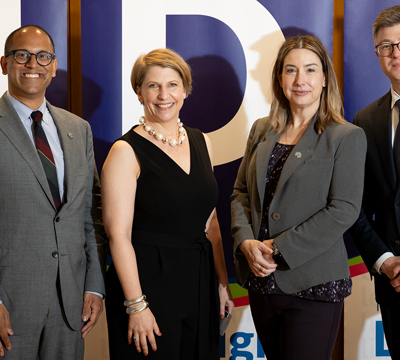 Photo of four people smiling and dressed in professional attire, standing in front of three banners spelling out UDL.