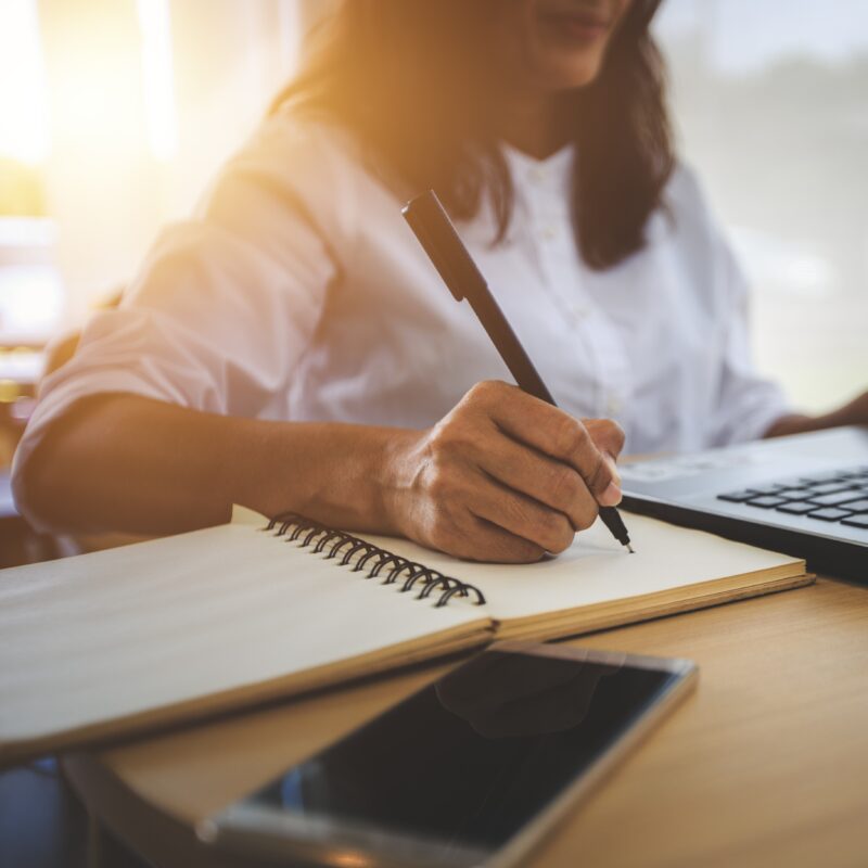 A person writing in a notebook at a desk with a laptop and phone