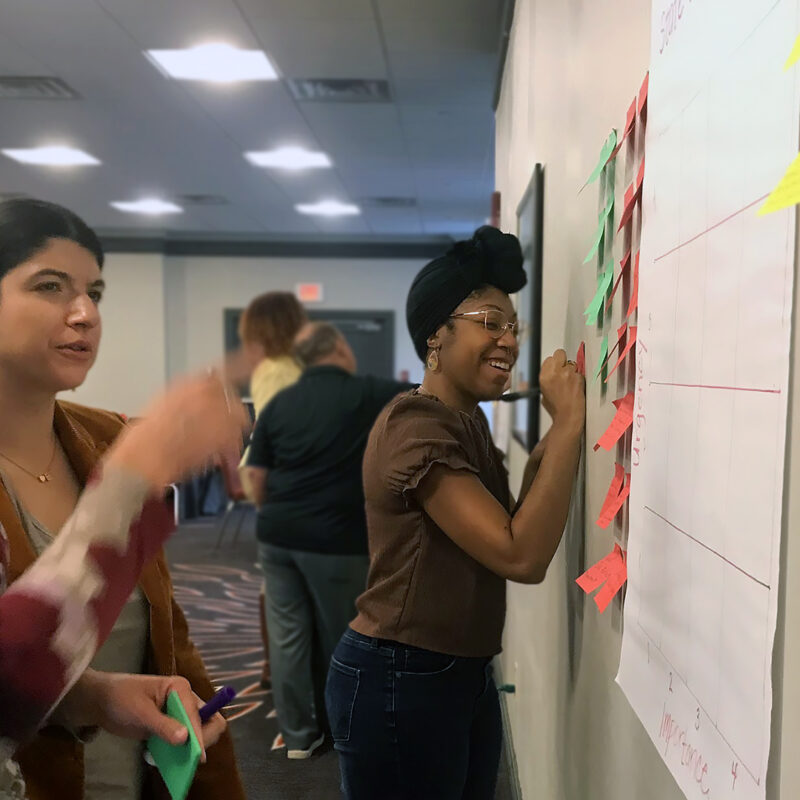 Photo of two adult women engaged in discussion pointing at a wall with sticky notes and chart paper. A woman in the background is smiling and writing on a sticky note on the wall., Illustration of a series of media representing varied ways of feeling, seeing and hearing content., Center on Inclusive Technology & Education Systems (CITES) logo, CAST mark, Various icons representing learning about UDL, Three CAST UDL Credentials badges