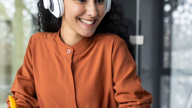 Woman in a conference room taking notes while wearing headphones and looking at her laptop