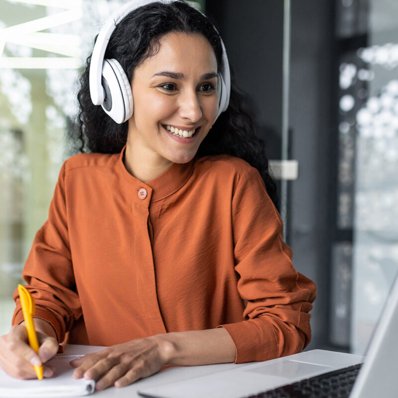 Woman in a conference room taking notes while wearing headphones and looking at her laptop