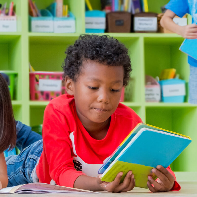 A bright classroom with colorful bins of books and supplies and a reading group of young students.